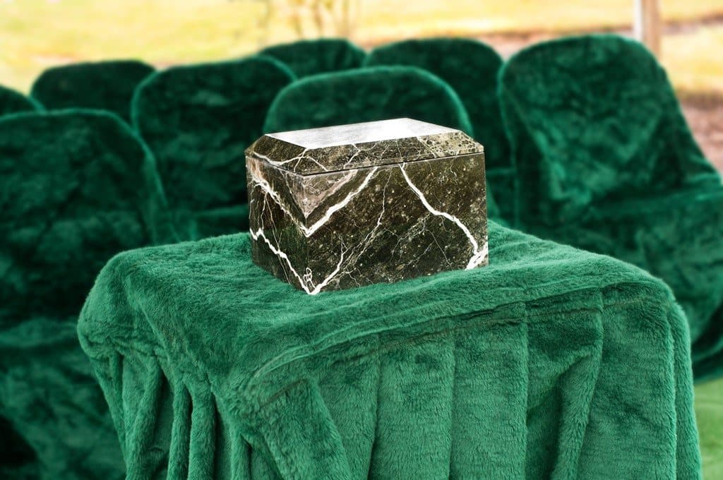 A marble-look urn on a table draped in green velvet at a graveside committal, with chairs behind it.
