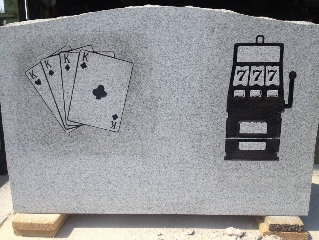 A gray granite stone etched with a hand of playing cards and a slot machine.