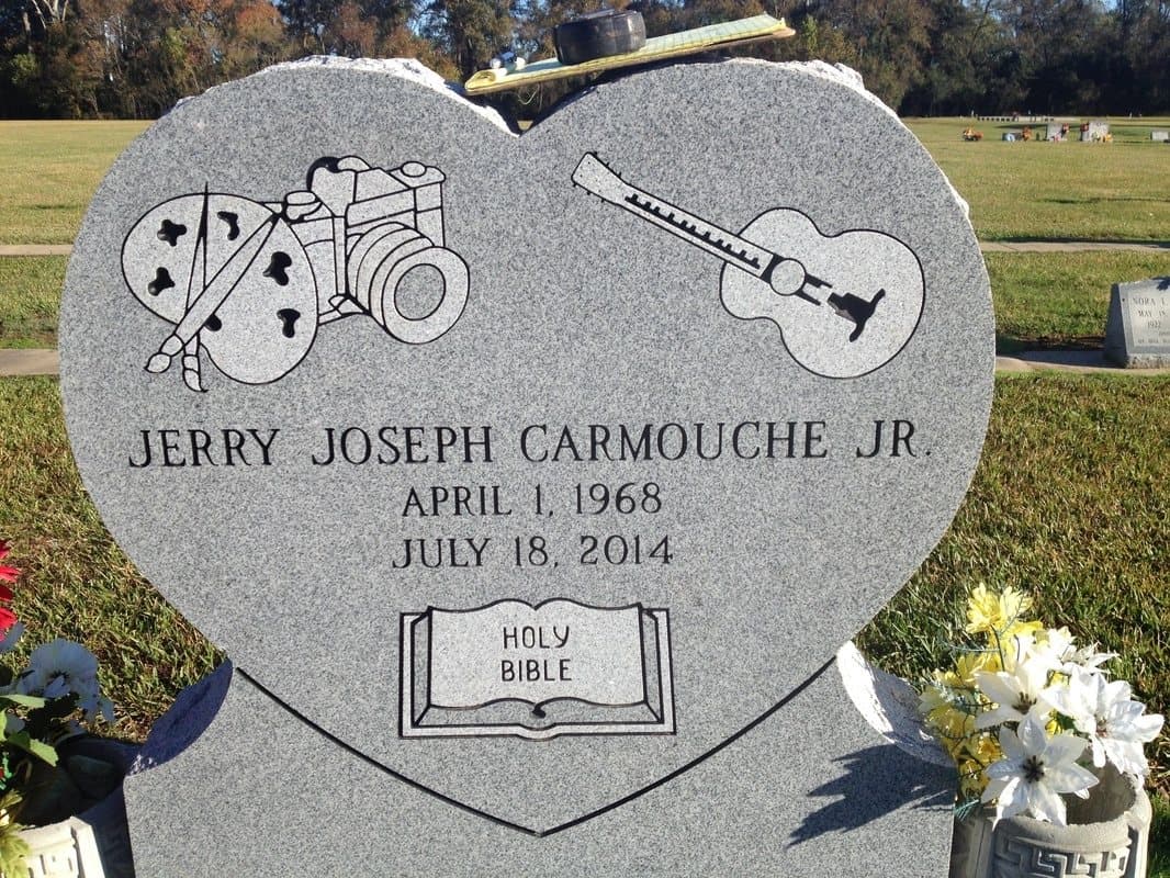 A heart-shaped gray granite headstone etched with a camera, a guitar, and an open Bible.