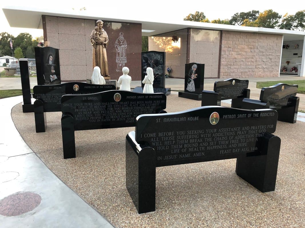 Black granite benches and monuments along a curved walk in a prayer garden, with a bronze statue of St. Anthony.