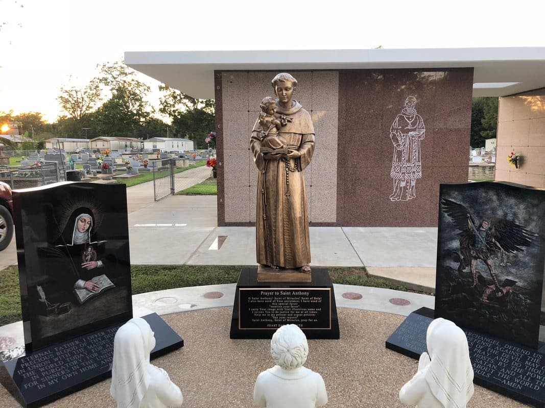 A bronze statue of St. Anthony on a black granite base, with etched monuments and kneeling statues around it.