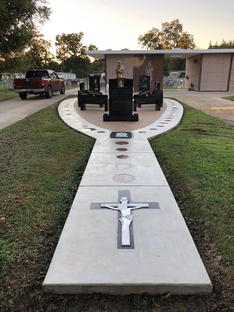 A long concrete walkway inlaid with a white crucifix, leading to granite benches and a statue.
