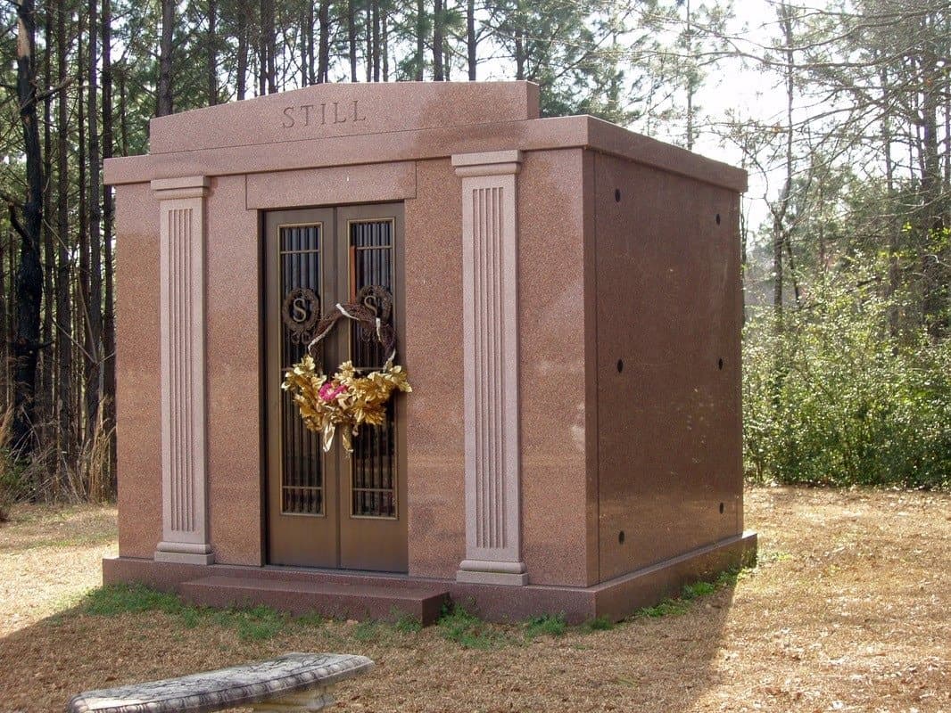 A red granite private mausoleum with columns and bronze doors, at the edge of a wooded cemetery.