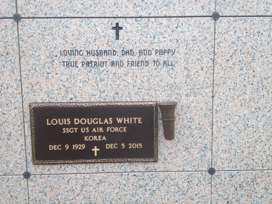 A veteran's bronze plaque on a pink granite crypt front, with a cross and a short tribute engraved above it.