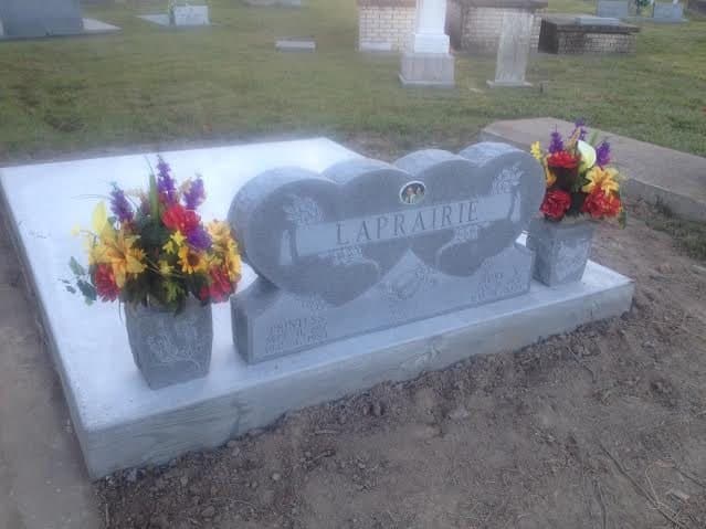 A heart-shaped double headstone on a new concrete slab, with flowers on each side.