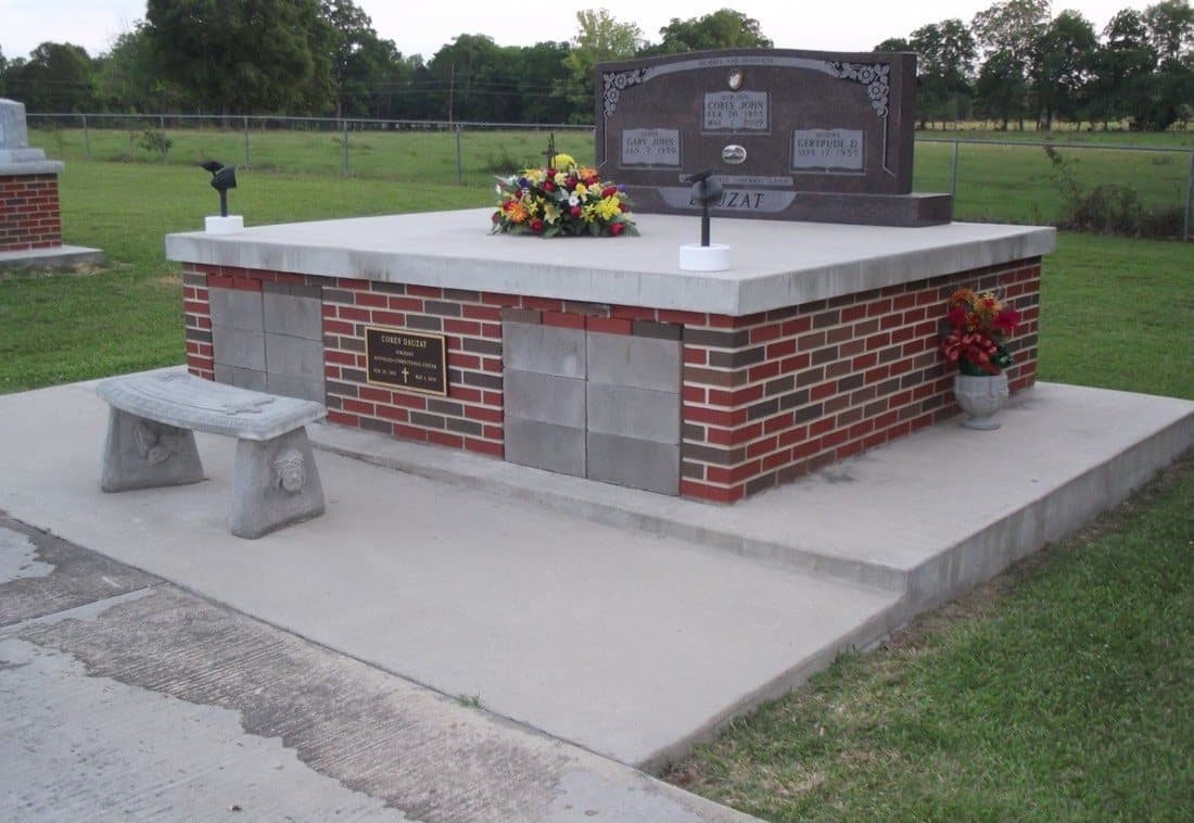 A red brick double tomb with bronze plaques, a granite headstone, and a bench on a wide concrete slab.