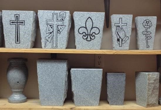 A shelf of granite vases etched with crosses, a fleur-de-lis, praying hands, and a rose.