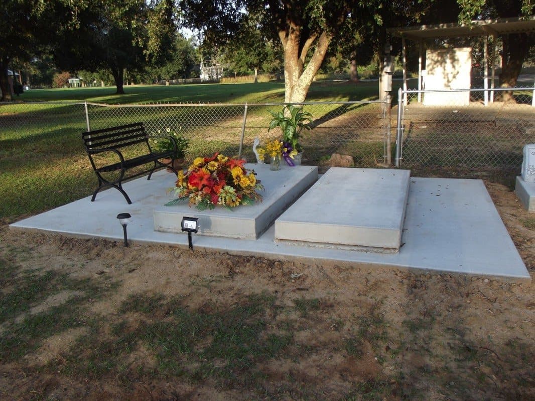 Two concrete vaults on a slab with a wide concrete apron, flowers, a bench, and a solar light.